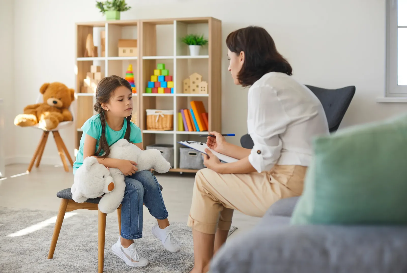 A therapist sitting on the floor beside a child who is holding a soft toy, listening attentively during a supportive session